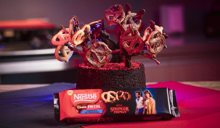 Bolo de chocolate decorado com galhos feitos de chocolate, formando uma árvore irregular onde ChocoPretzels ficam pendurados como pequenos frutos.