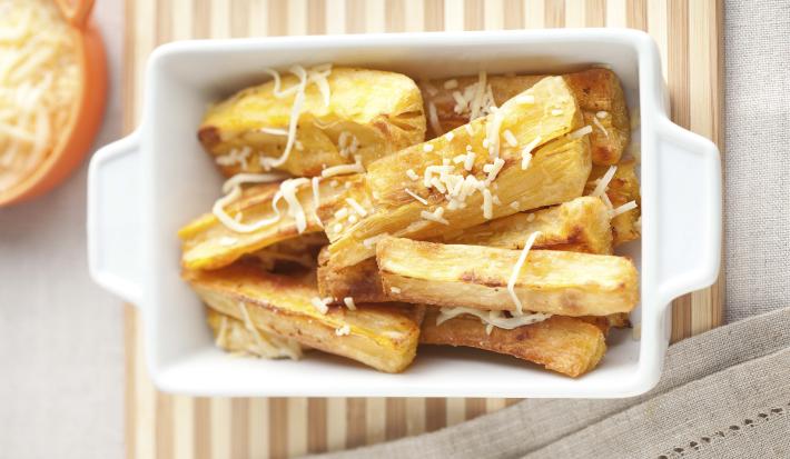 Fotografia em tons de bege em uma mesa de madeira com uma toalha bege e uma travessa retangular branca com as mandiocas assadas com parmesão dentro.