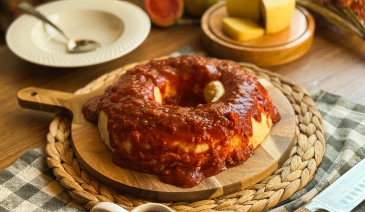 Fotografia de um pudim de queijo colonial com calda de goiabada sobre uma tábua de madeira redonda. Ao fundo tem um prato branco fundo, colher de sopa e mini tábua grossa de madeira com o queijo colonial cortado.