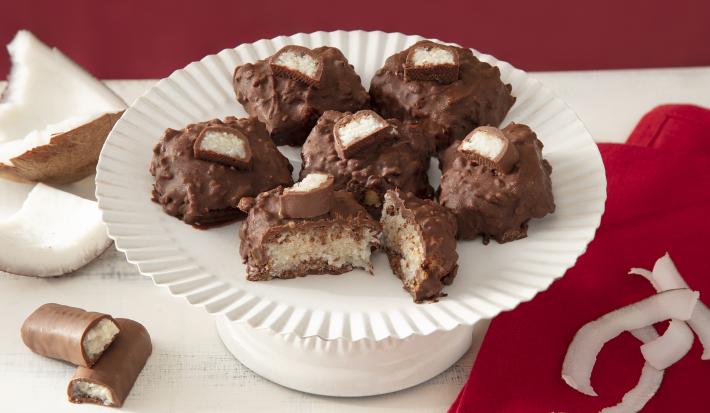 Foto da receita de cocada de prestígio. Observa-se uma boleira branca com 6 cocadas banhadas no chocolate. A da frente está cortada e é possível ver o coco de dentro