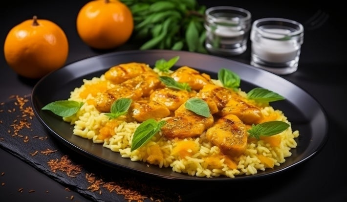 Em cima de uma mesa, prato redondo preto om arroz cremoso amarelo com pedaços de frango e decorado com folhas de hortelã.