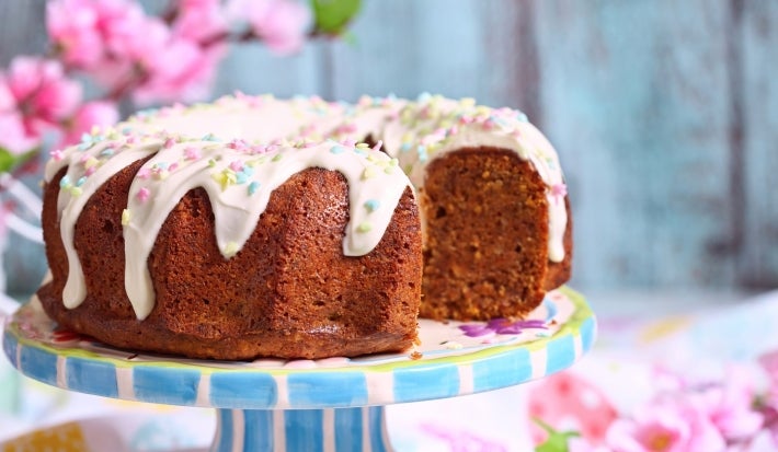 Fotografia de um bolo bundt elegantemente decorado, coberto com glacê branco e confeitos em tons pastel de estrelas e bolinhas. O bolo, de cor dourada e fofinho, está em um suporte listrado azul e branco, com um fundo de flores de cerejeira.