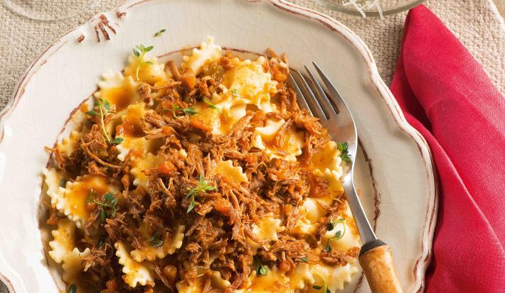 Fotografia em tons de bege em uma mesa de madeira com um guardanapo de pano vermelho ao lado, um prato redondo fundo branco com detalhes vermelhos e o farfalle com ragu de carne dentro dele.