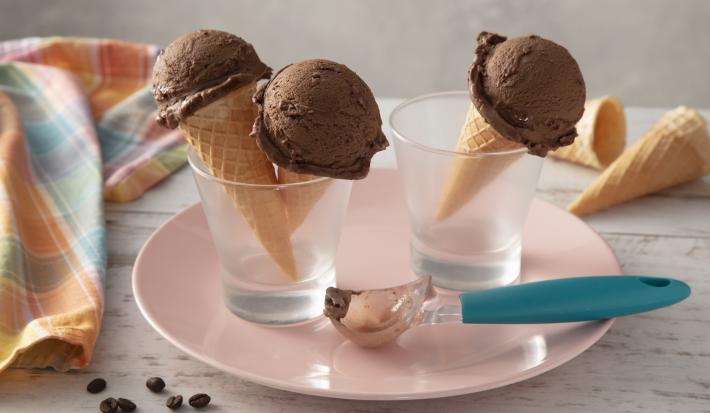 Fotografia em tons de azul e rosa de uma bancada, ao centro um prato rosa com dois copos, dentro deles casquinhas de sorvetes com uma bola do sorvete de café com chocolate. Sobre o prato, um boleador de sorvete com cabo azul. Fotografia em tons de azul e rosa de uma bancada, ao centro um prato rosa com dois copos, dentro deles casquinhas de sorvetes com uma bola do sorvete de café com chocolate. Sobre o prato, um boleador de sorvete com cabo azul.