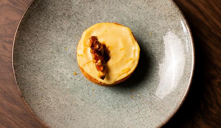 Fotografia em tons de marrom com um prato cinza ao centro. Dentro do prato existe um pequeno bolo de massa branca recheado com um creme de cor amarelada e coberto com castanhas caramelizadas.