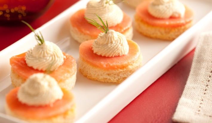Fotografia em tons de vermelho em uma bancada de madeira com um jogo americano vermelho, um recipiente branco retangular com alguns canapés de salmão em cima dele.