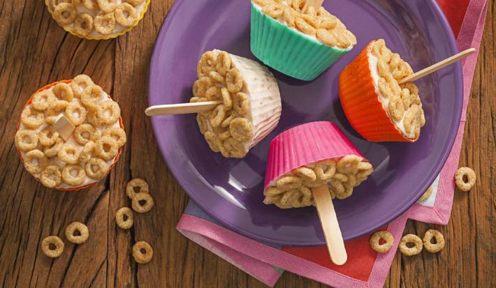 fotografia tirada de uma bancada marrom que contém um prato redondo roxo com 4 forminhas nas cores rosa, vermelho, branco e verde e todas contém um geladinho coberto com cereal e um palito de sorvete ao centro de cada um.