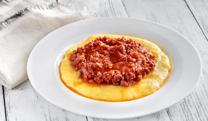 fotografia em tons de branco, amarelo e vermelho de uma bancada branca vista de cima. Contém um prato redondo branco com polenta, e por cima um molho bolonhesa.