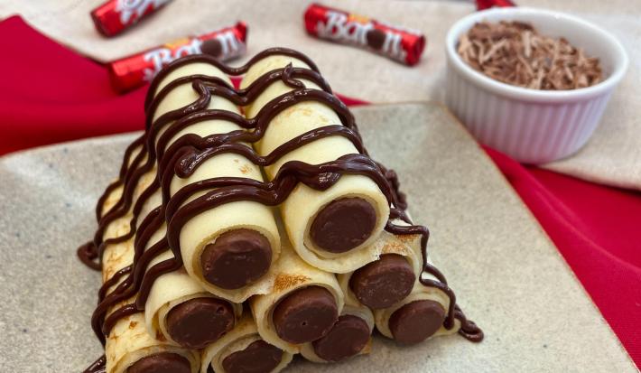 Fotografia em tons de vermelho com uma torre de crepes recheados ao centro. Os crepes encontram-se empilhados e recheados com chocolate Baton ao Leite e cobertos com uma calda de chocolate.