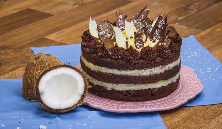 Foto da receita de Bolo Naked de Prestígio, decorado com pedaços de chocolate, sobre um prato rosa, numa bancada de madeira com tecidos azuis e dois pedaços de coco fresco aberto