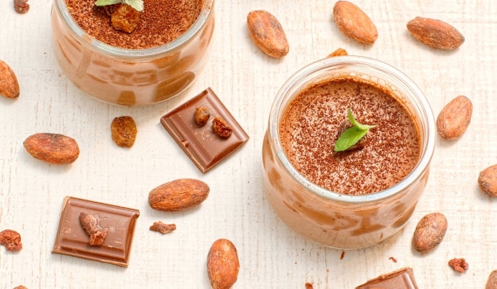 Fotografia em tons claros de quatro recipientes de vidro pequenos com a sobremesa, finalizados com a ganache, chocolate em pó, folhas de hortelã, e algumas castanhas. Os recipientes estão sobre uma mesa de tom claro, com castanhas e cacau espalhados