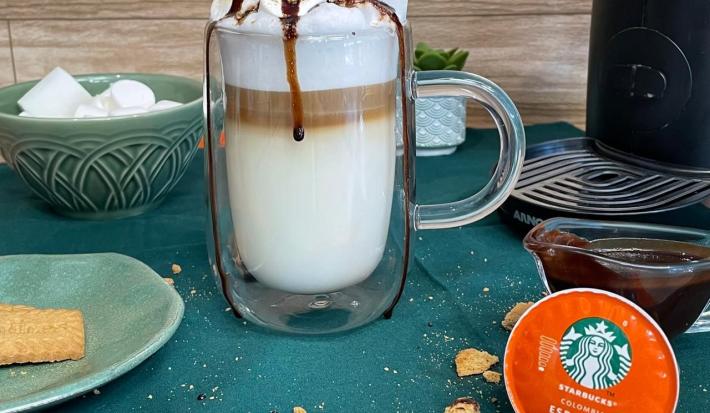 Fotografia em tons de verde em uma bancada com um pano verde, uma xícara de vidro grande com a bebida de leite e café preparada dentro dela. Decorada com marshmallow, calda de chocolate e biscoito triturado.