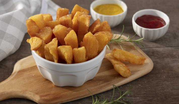 Fotografia em tons de amarelo em uma bancada de madeira escura, uma tábua de madeira, um pote branco fundo com várias mandiocas fritas. Ao fundo, um pano bege xadrez e dois potinhos um com ketchup e um de mostarda.