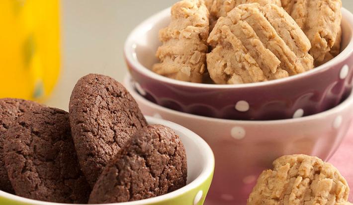 Fotografia em tons de rosa e bege em uma bancada de madeira clara, um paninho rosa, três potinhos coloridos, verde, rosa e roxo, com bolinhas brancas e os biscoitinhos de chocolate e de aveia dentro deles. Ao fundo, uma jarra com suco de laranja.