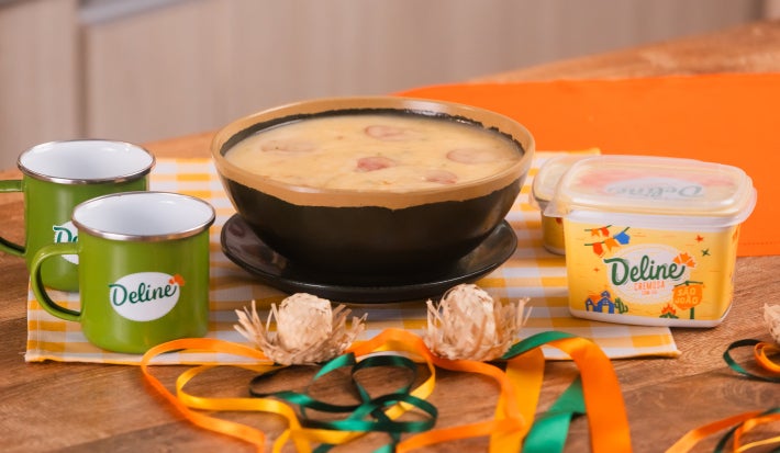 Caldo cremoso de macaxeira com rodelas de calabresa em tigela, acompanhado de manteiga Deline e canecas para festa junina