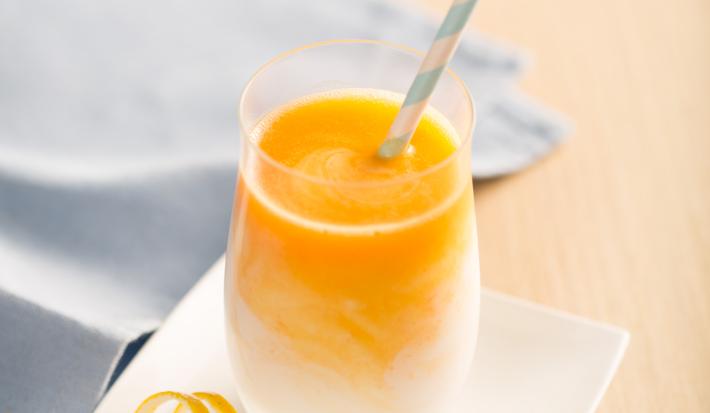 Fotografia em tons de laranja e azul em uma bancada de madeira clara, um pano azul, um prato pequeno branco quadrado com um copo de vidro em cima dele e o smoothie de laranja, manga e cenoura dentro dele.