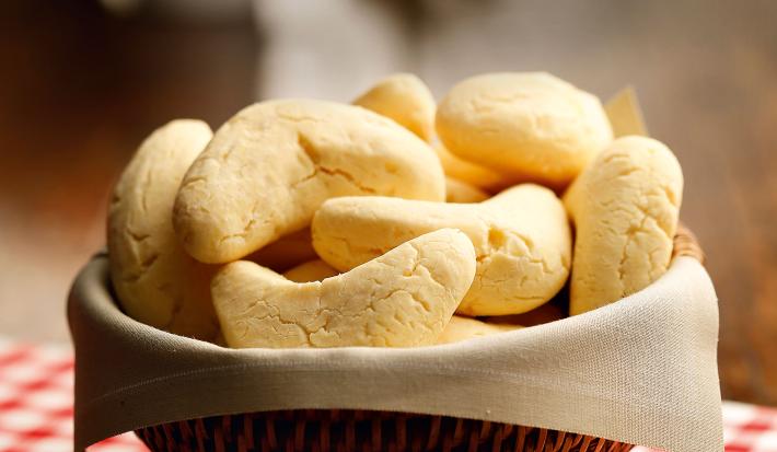 Fotografia em tons de vermelho em uma bancada de madeira, um pano vermelho xadrez, uma cesta de vime marrom, um guardanapo branco de pano com as chipas, pães à base de ovos, manteiga e queijo, dentro dela.