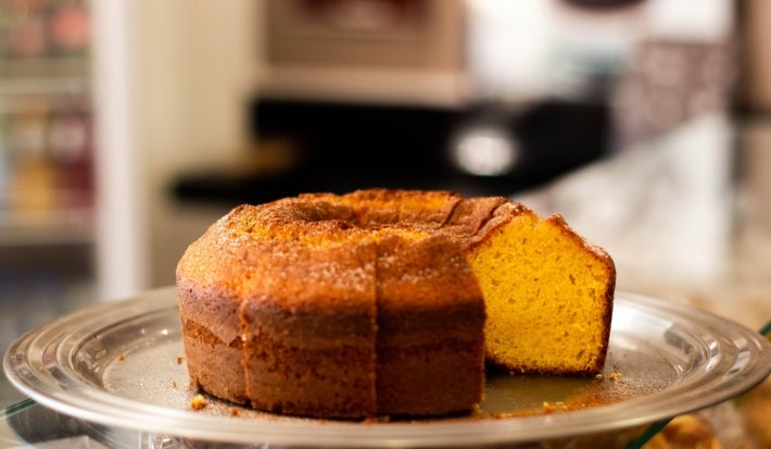 Um bolo de cenoura redondo com furo no meio está parcialmente cortado e exposto sobre um prato de metal. A textura está fofa e dourada por dentro, com uma casquinha crocante por fora.