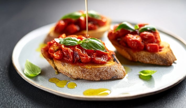 Fotografia em tons claros de três fatias de pão italiano torrado sobre um prato raso branco, apoiado em uma mesa preta. Sobre as torradas tem um pouco de confit de tomates cereja com folhas de manjericão e azeite.