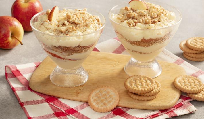 Fotografia em tons de cinza, vermelho e branco de uma bancada cinza com um paninho xadrez branco e vermelho com uma tábua de madeira, sobre ela duas taças com pavê de maçã e biscoitos tostines espalhados pela mesa. Ao fundo duas maçãs.
