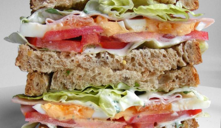 Fotografia de um sanduíche em camadas no pão de forma integral com tomate, repolho, maionese, presuntada e alface. O sanduíche está cortado na metade, sendo uma em cima da outra, que está sobre uma mesa branca.