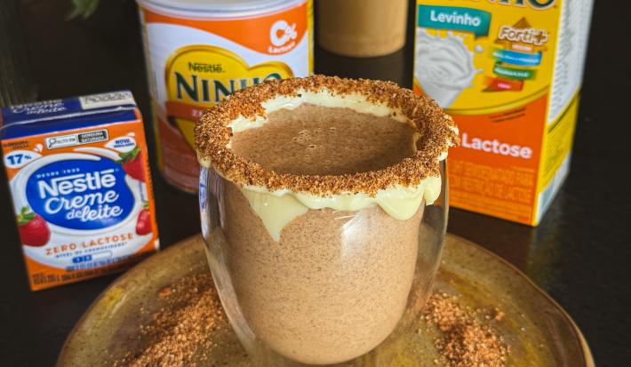 Fotografia de uma xícara de vidro com uma bebida de ninho com o creme de ninho na borda e ninho tostado também na borda e no prato raso em que a xícara está apoiada. O prato está sobre uma mesa preta, ao lado do prato tem produtos zero lactose nestlé