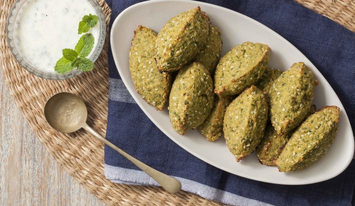 Fotografia em tons de verde, azul e branco, ao centro uma travessa branca com os bolinhos de falafel, ao lado para acompanhar um potinho de vidro com o molho de iogurte e uma folhinhas verdes para decorar. Abaixo está um guardanapo azul.