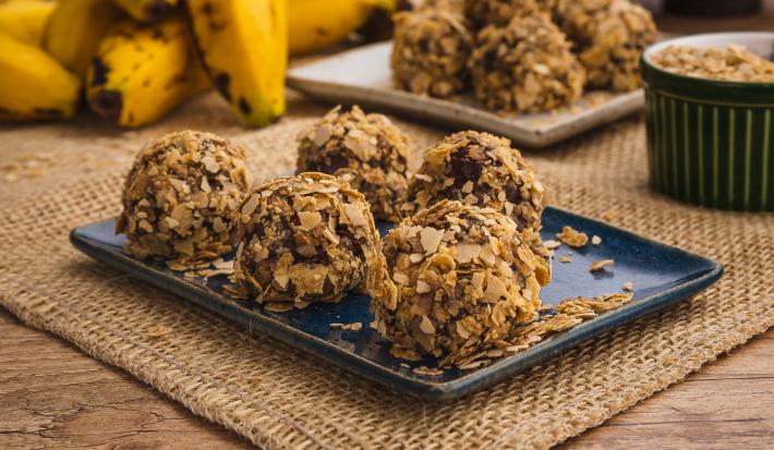 Fotografia em tons de amarelo e azul em uma bancada de madeira, um jogo americano de palha, um recipiente retangular azul com alguns brigadeiros de banana coberto com cereais Neston. Ao fundo, um cacho de banana.