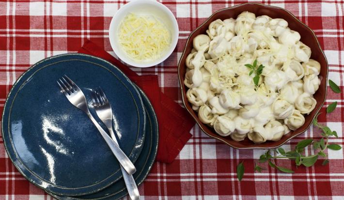 Fotografia em tons de azul, vermelho e branco com dois pratos azuis e garfos sobre guardanapo vermelho, refratário com massa ao creme com potinho contendo queijo ralado ao alado, tudo sobre toalha branca com quadriculados em tons de vermelho. Fotografia em tons de azul, vermelho e branco com dois pratos azuis e garfos sobre guardanapo vermelho, refratário com massa ao creme com potinho contendo queijo ralado ao alado, tudo sobre toalha branca com quadriculados em tons de vermelho.