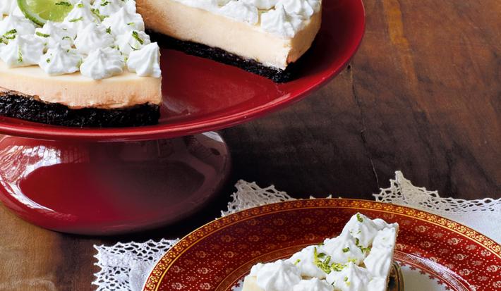 Fotografia em tons de vermelho e branco, com boleira vermelha contendo uma torta com creme e cobertura brancos, ao lado prato branco com borda vermelha com um pedaço de torta sob uma espátula e sobre guardanapo branco, tudo sobre bancada de madeira.