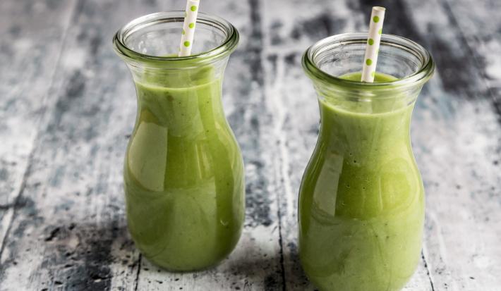Fotografia de duas garrafas de vidro pequenas com um refresco de salsão, e em cada garrafinha há um canudo branco com bolinhas verdes. Os refrescos estão sobre uma mesa de madeira branca com detalhes pretos.