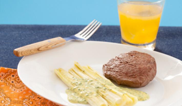 fotografia em tons de branco, azul e laranja de uma bancada vista de frente. Contém um pano laranja, um prato redondo e branco com o alho-poró e um medalhão de filé mignon e ao lado um garfo. Ao fundo um copo transparente com uma bebida laranja