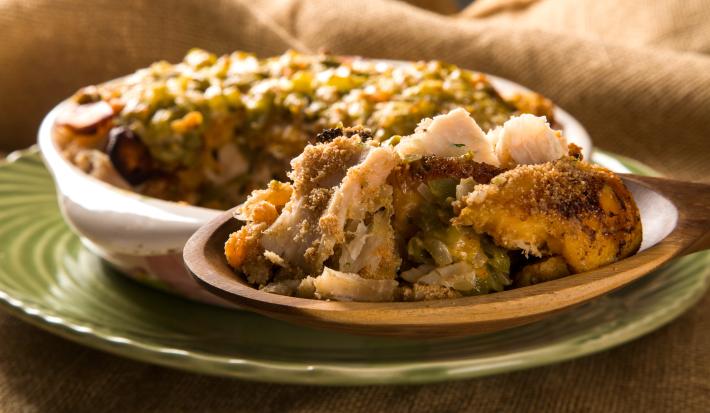 Foto da receita de Pirarucu Assado. Observa-se o peixe disposto com os legumes e a banana em um recipiente branco e, no plano principal, observa-se uma colher de pau contendo a preparação de perto.