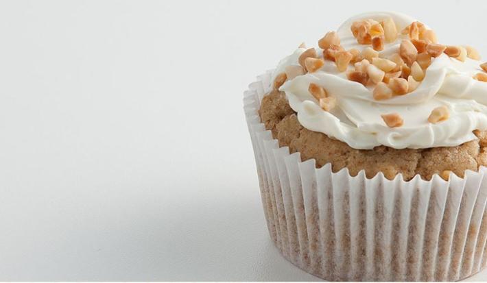 Fotografia em tons de bege em uma mesa branca com uma forminha de doce grande branca com o cupcake de massa integral dentro dele. Decorado com castanhas-de-caju.