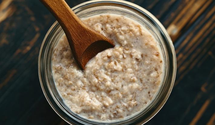 Fotografia vista de cima de um recipiente de vidro decorativo com uma receita de overnight oats de coco. O recipiente está sobre uma mesa de madeira.