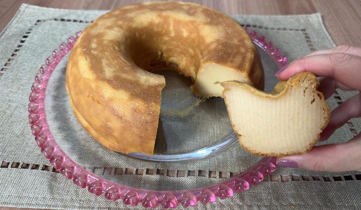 Fotografia de um bolo de farinha láctea sobre um apoio decorativo de vidro raso apoiado em um pano de tom verde.