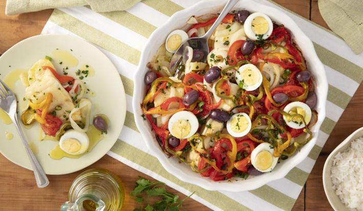 Imagem vista de cima de uma travessa redonda com bacalhoada com rodelas de ovos, pimentões, azeitonas e tomate. Ao lado, um prato branco com uma porção de bacalhoada, um vidro de azeite e um bowl de arroz branco.