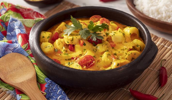 Fotografia em tons de amarelo em uma bancada de madeira de cor branca. Ao centro, uma panela preta contendo a moqueca. Ao lado, há um pano florido vermelho com uma colher de pau em cima. Ao fundo, há uma tigela de arroz.