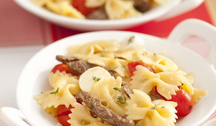 Fotografia em tons de vermelho em uma bancada de madeira, um jogo americano vermelho e branco, um prato branco redondo com o macarrão com tirinhas de carne e tomate.