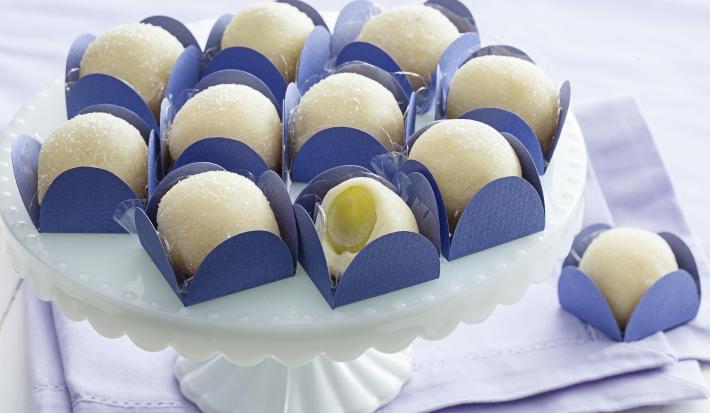 Fotografia em tons de branco e azul de uma bancada branca com paninho azul, sobre ele uma bailarina branca com docinhos de brigadeiro branco com uva verde dentro de forminhas quadradas azuis.