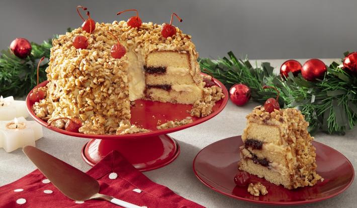 Foto com decoração natalina da receita de bolo de coroa de frankfurt servida sobre uma bailarina vermelha com um pedaço cortado ao lado sobre um prato de porcelana vermelho. Tudo isso em cima de uma mesa branca