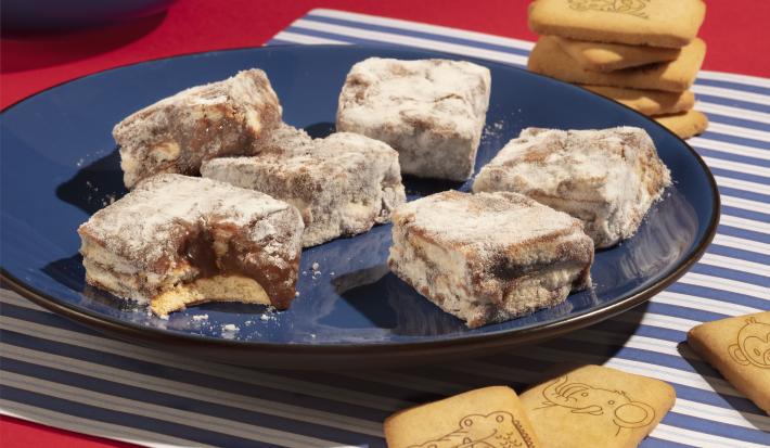 Fotografia em tons de azul e vermelho de uma bancada vermelha com paninho listrado de azul e branco, sobre ele um prato azul escuro com palha italiana de biscoito passatempo. Ao fundo biscoitos passatempo leite espalhados.