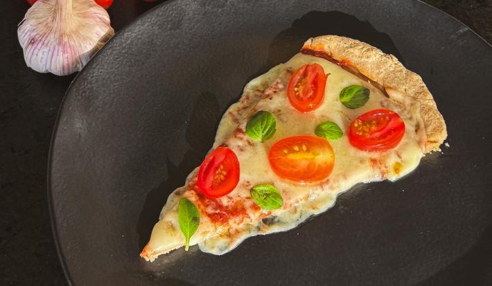 Fotografia de um prato raso preto sobre uma bancada preta. Dentro do prato tem uma fatia de pizza com queijo, tomate e manjericão. Ao lado do prato tem duas cabeças de alho inteiras com vários tomates cereja e um vidro de azeite.