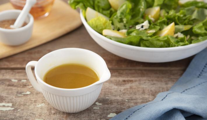 Fotografia em tons de azul em uma bancada de madeira escura, um pano azul, uma tábua de madeira, um potinho branco com o molho de mostarda e mel dentro. Ao fundo, um recipiente com salada verde para acompanhar o molho e uma tábua com mais potinhos.