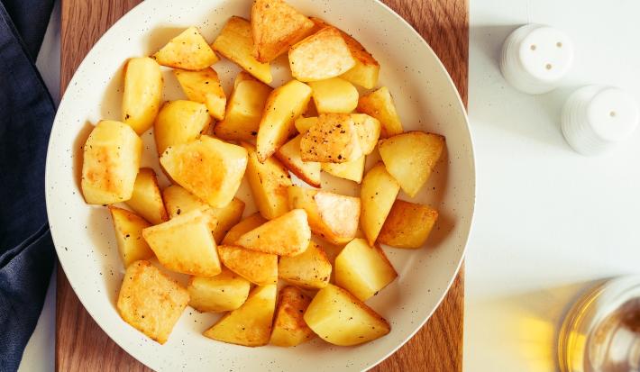 fotografia em tons de amarelo e branco de uma bancada branca vista de cima. Contém uma tábua de madeira marrom, por cima um recipiente redondo branco e dentro batatas picadas.