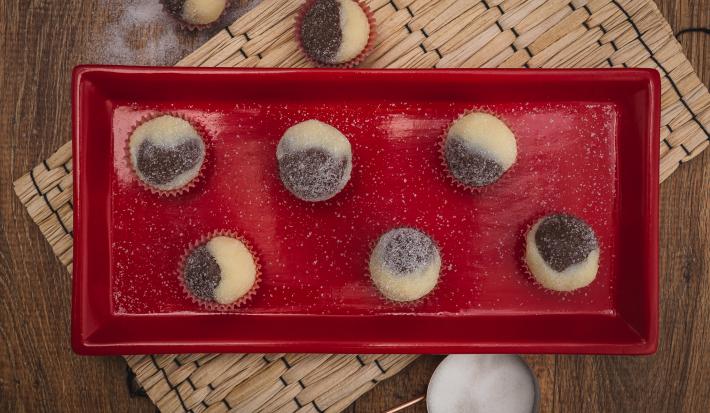 Fotografia em tons de vermelho em uma bancada de madeira escura, um jogo americano de palha, um recipiente retangular vermelho com brigadeiros mesclados de brigadeiro branco e preto em forminhas de vermelho.