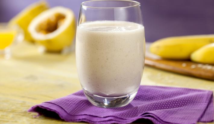 Fotografia em tons de roxo, pardo, branco e amarelo de uma bancada vista de frente. Contém um pano roxo com um copo transparente por cima com a vitamina. Ao junto maracujá cortado e bananas