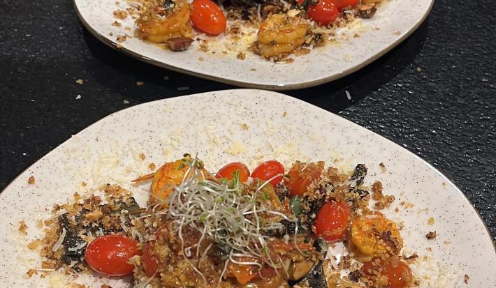 Fotografia de dois pratos de vidro parcialmente quadrados, de pontas arredondadas, sobre uma mesa preta. Dentro dos pratos tem uma massa linguine nero com molho rústico de tomate grape, camarões e farofa de castanhas.