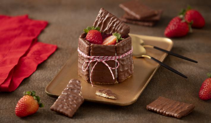 Foto de uma bancada escura com alguns tecidos vermelhos, morangos e biscoitos cobertos de chocolate espalhados. Ao centro há um prato retangular dourado com a receita pronta montada e decorada, acompanhada de duas colheres douradas e pretas