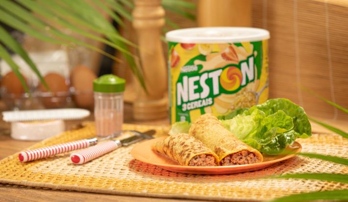 Foto de uma bancada, sobre ela há um jogo americano e nele um prato com dois wraps recheados. Ao fundo há uma lata de Neston 3 Cereais, alguns talheres e outros utensílios. Há algumas plantas compondo a imagem.